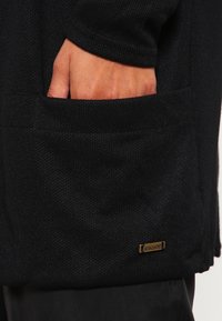 Svart stickad jacka med ett texturerat mönster, som har en sidoficka och en liten guldfärgad logotyp. Materialet verkar mjukt och varmt.