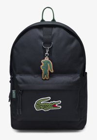 Schwarzer Rucksack mit einer Vordertasche, versehen mit einem grünen und weißen gestickten Logo und einem grünen Schlüsselanhänger in Form eines Krokodils.