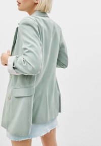 Světle zelená blazer s uvolněným střihem, se švy na ramenou, dvěma knoflíky a jednou přední kapsou. Bílé manžety vykukují z ohrnutých rukávů.