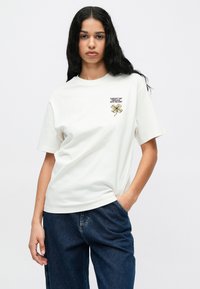 Biela bavlnená tričko s voľným strihom a krátkymi rukávmi, s menším vyšitým žltým a čiernym vážkou na ľavej strane hrudníka.