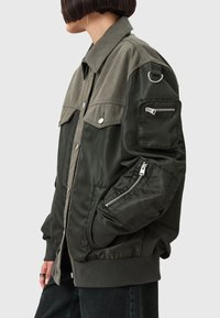 Giacca bomber verde con un mix di tessuto opaco e lucido, vestibilità oversize, chiusura con bottoni e molteplici tasche con zip sulle maniche.