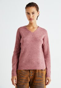 Rosa, gerippter Strickpullover mit V-Ausschnitt und langen Ärmeln, mit lockerem Schnitt. Kombiniert mit braunen, gemusterten hochtaillierten Hosen.