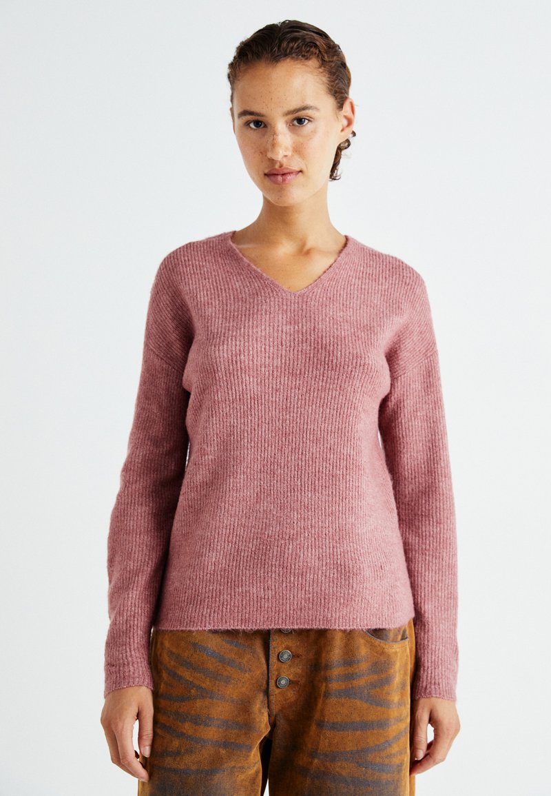 Rosa, gerippter Strickpullover mit V-Ausschnitt und langen Ärmeln, mit lockerem Schnitt. Kombiniert mit braunen, gemusterten hochtaillierten Hosen.