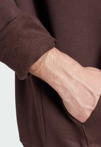 Sweat-shirt marron avec une texture côtelée et des poignets tissés, montrant une main reposant sur la poche, mettant en valeur son design décontracté.