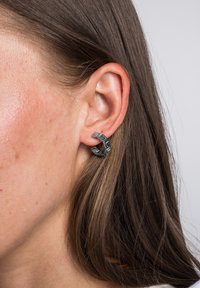 Pendientes de aro metálico gris con forma circular, que presentan un patrón geométrico y una superficie texturizada, llevados en el lóbulo.