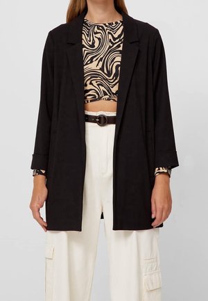 Mujer con blazer negro sobre un top corto de estampado abstracto beige y negro, combinado con pantalones cargo blancos de talle alto y un cinturón negro.