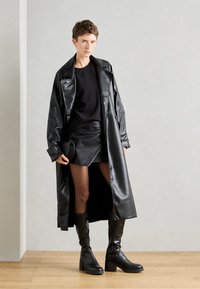 Trench-coat en cuir noir avec une finition brillante, porté sur un haut noir et une mini-jupe en cuir. Des bottes hautes noires à talons complètent la tenue.