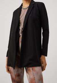 Svart kavaj med långa ärmar, skräddarsydd design och en slät yta, matchad med en ljusbrun topp och knytframtill, bärs med mönstrade leggings.