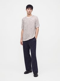 Jeune homme portant une chemise légère à manches courtes texturée et un pantalon large sombre à motifs avec des chaussures noires, debout devant un fond uni.