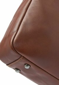 Ecke einer braunen Ledertasche mit glatter Textur, sichtbaren Nähten und Metallbeschlägen. Abgerundete Kanten und ein poliertes Finish.