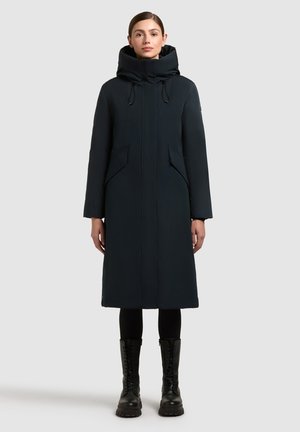 Nő hosszú fekete párnázott télikabátban, magas gallérral és kapucnival, fekete leggingsszel és fekete fűzős csizmával kombinálva, egyenesen áll.