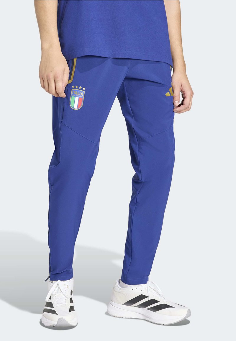 Niebieskie spodnie sportowe o zwężanym kroju, z odznaką Włoch, żółtymi akcentami i kieszeniami na zamek. Noszone z białymi sneakersami, czarne paski.