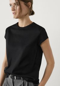 Mladá žena s světle hnědými vlasy vybranými do nízkého drdolu, oblečená v černém tričku s krátkými rukávy, které má zasunuté do šedých kalhot s černým páskem.