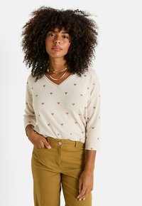 Pull beige à col en V avec de petits motifs en forme de cœur marron, manches trois-quarts et tissu texturé. Associé à un pantalon taille haute olive.