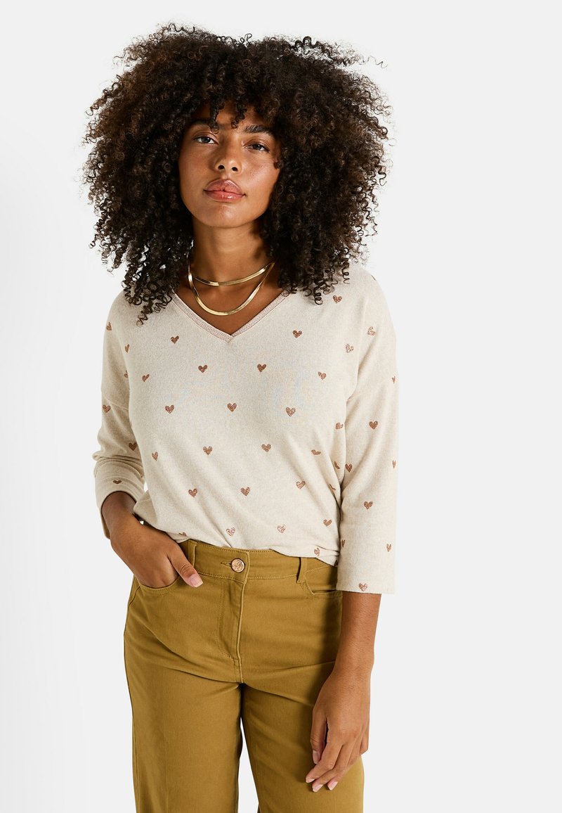 Pull beige à col en V avec de petits motifs en forme de cœur marron, manches trois-quarts et tissu texturé. Associé à un pantalon taille haute olive.