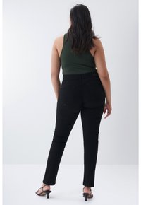 Femme aux longs cheveux foncés portant un haut sans manches vert foncé, un pantalon noir ajusté et des sandales à talons noires, debout de dos à la caméra.