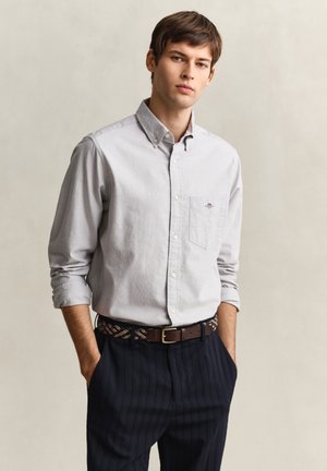 Junger Mann mit braunen Haaren, der ein hellgraues Hemd mit Knöpfen trägt, dunkel gestreifte Hosen, gemusterten Gürtel, steht mit den Händen in den Taschen.