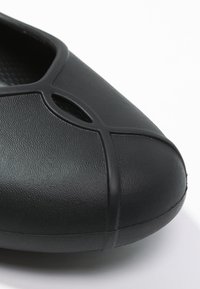 Chaussure en caoutchouc noire avec un bout arrondi, présentant une surface texturée et un détail découpé décoratif sur le dessus.