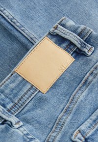Tissu en denim bleu clair avec double couture, passants pour ceinture et une étiquette en cuir beige vierge cousue sur la ceinture.