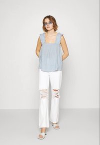 Top sem mangas azul claro com bordas com babados, combinado com calças brancas de perna larga desgastadas. Modelo usa óculos de sol azuis e sandálias de salto cinza claro.