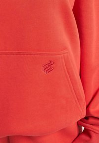 Rød hettegenser med en myk tekstur, som har en frontlomme og en brodert logo i en matchende nyanse. Enkel, uformell design.