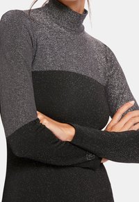 Femme portant une robe noire et argentée pailletée à manches longues et col montant, les bras croisés sur sa poitrine.