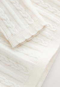 Tissu tricoté blanc cassé avec un motif torsadé et des bords côtelés, offrant une texture douce et un détail d'ourlet festonné.