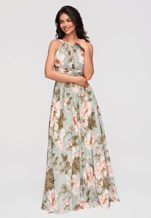 Femme aux cheveux foncés portant une robe fleurie pastel sans manches, longue jusqu'au sol, souriante et posant sur un fond clair uni.