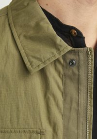 Olivgrön jacka med strukturerad krage, dragkedjestängning och knappdetalj. Tyget verkar lätt och smidigt med en subtil textur.