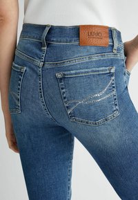 Modri jeans iz denima s srednje visokim pasom, z rjavim usnjenim oznakami in svetlečimi kamenčki na zadnjih žepih.
