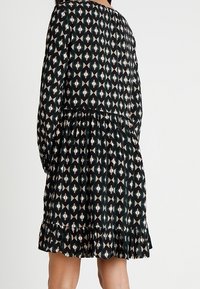 Černé šaty s geometrickým vzorem v krémové, růžové a tyrkysové barvě. Dlouhé rukávy, nařasený pas a volánkový spodní lem. Měkká textilie.