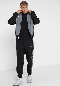 Tracksuit Nike preto e cinza com jaqueta de fecho éclair e calças a condizer com riscas brancas laterais. Combinado com ténis desportivos cinzentos.