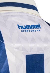 Dos d'un maillot de sport blanc avec motif géométrique bleu, logo Hummel Sportswear bleu et bande de col marine.