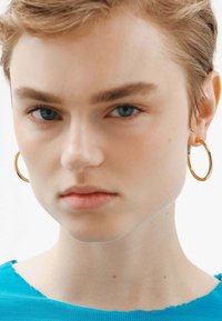 Aros de oro en una modelo con cabello corto, que lleva una camisa azul texturizada. Los aros tienen un acabado suave y pulido.