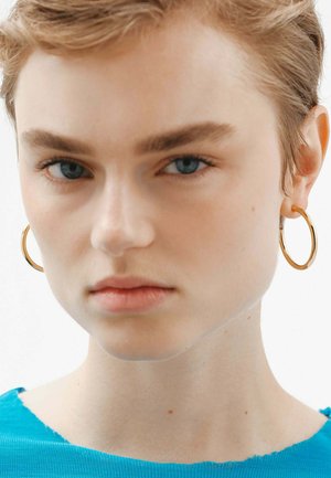 Aros de oro en una modelo con cabello corto, que lleva una camisa azul texturizada. Los aros tienen un acabado suave y pulido.