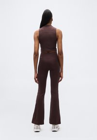 Γυναίκα που στέκεται με την πλάτη προς την κάμερα, φοράει σκούρο καφέ αμάνικο τοπ και ταιριαστά παντελόνια καμπάνα με λευκά αθλητικά παπούτσια.