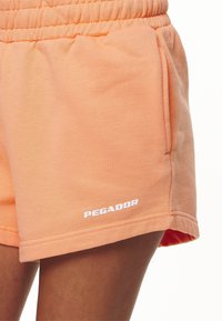 Krémové šortky z mäkkej tkaniny, s elastickým pásom, bočnými vreckami a bielym logom "PEGADOR" na prednej strane.