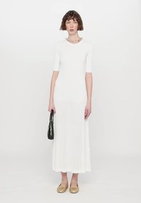 Robe maxi blanche à rayures, avec manches courtes, présentant un haut ajusté et un bas évasé. Le modèle tient un sac à main noir et porte des ballerines dorées.