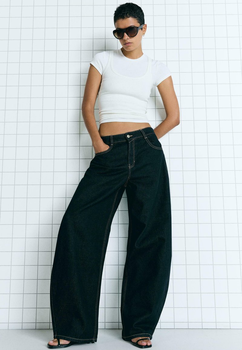 Wide-leg črne kavbojke z oranžnim šivom, v kombinaciji z belim prilegajočim vrhom. Model nosi temna sončna očala, roke ima v žepih, pred mrežasto ozadjem.