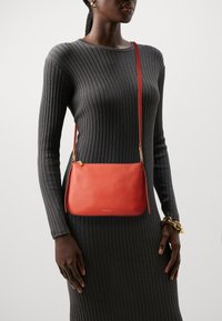 Kobieta ubrana w dopasowaną, ciemnoszaro prążkowaną sukienkę, pomarańczową torebkę na ramię, złote kolczyki w kształcie kół oraz masywną złotą bransoletkę na prawej nadgarstku.