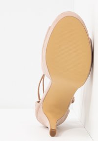 Chaussure à talon haut en daim beige avec un talon fin, semelle vers l'avant, positionnée verticalement contre une surface blanche.