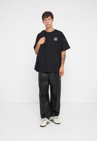 Svart oversized t-skjorte med grønn flammegrafikk på brystet, kombinert med svarte skinnbukser og hvite sneakers.