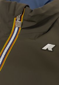 Giacca marrone con una cerniera argentata e un accento a strisce multicolori. Ha un patch con logo sul petto, realizzata in materiale liscio.