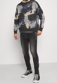 Suéter gris con patrones abstractos en azul, negro y beige; cuello redondo; combinado con jeans oscuros y zapatillas negras con detalles blancos.