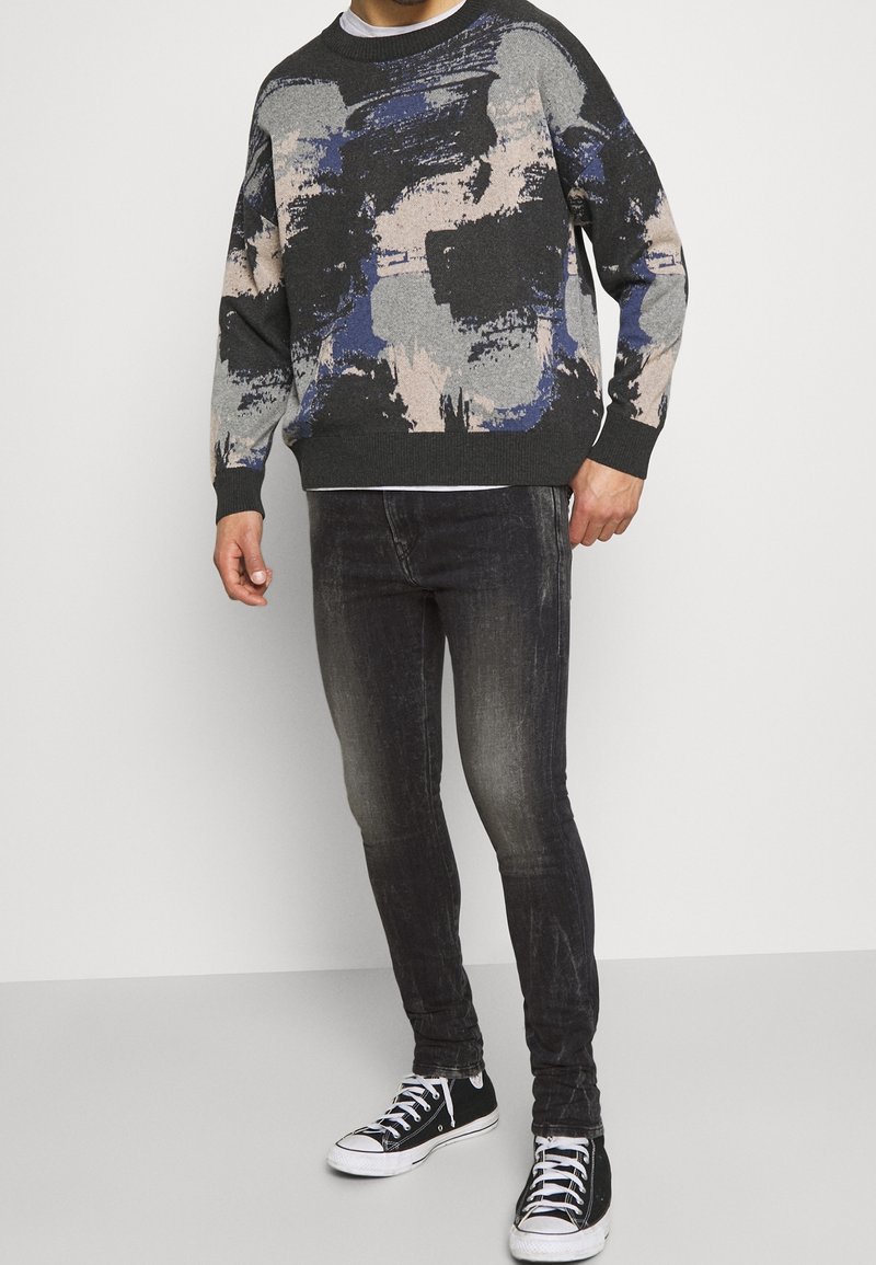 Suéter gris con patrones abstractos en azul, negro y beige; cuello redondo; combinado con jeans oscuros y zapatillas negras con detalles blancos.