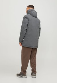 Grå vandtæt jakke med hætte, der har et strømlinet design og isolerende stof, parret med brune afslappede bukser og grå sneakers.