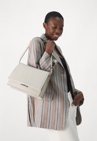Beige strukturert håndveske med klaffelukking, glatt tekstur og detaljer i sølvmetall, holdt av en person som har på seg en stripete blazer.