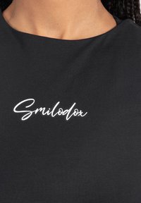 Primo piano del collo di una maglietta nera con la scritta corsiva bianca "Smilodox" centrata sul petto, in contrasto con uno sfondo di pelle marrone.