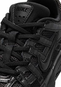 Κοντινό πλάνο μαύρου αθλητικού παπουτσιού Nike που δείχνει υφασμάτινο πλέγμα, δερμάτινες επενδύσεις, κορδόνια σε σταυρωτό μοτίβο και ανάγλυφο λογότυπο Nike στη γλώσσα.