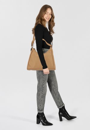 Hellbraune Schultertasche mit dreieckiger Form, strukturierter Oberfläche und verstellbarem Trageriemen. Kombiniert mit grauen Jeans und schwarzen, hohen Stiefeletten.